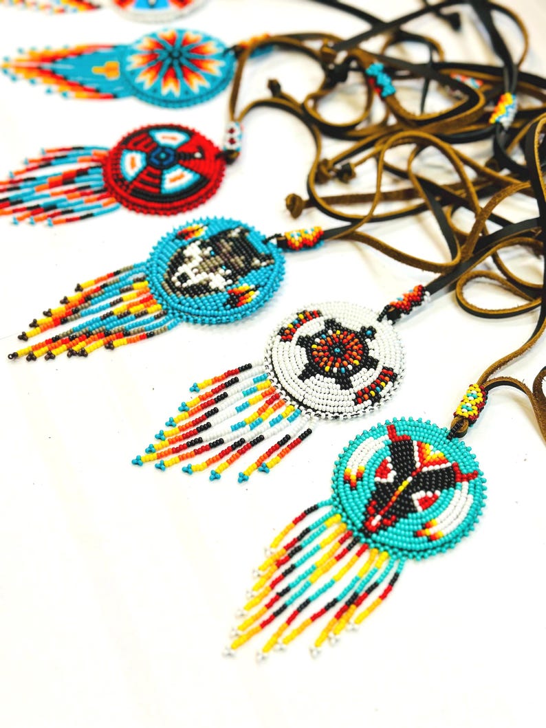 May include: A collection of beaded necklaces with colourful fringe. Each necklace features a round pendant with a different design, including a wolf, a turtle, and a geometric pattern. The necklaces are made with brown leather cords.