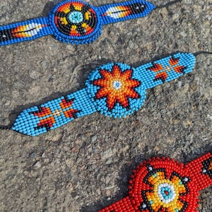 May include: Three beaded bracelets with colorful geometric patterns. The bracelets are made with blue, red, orange, yellow, white, and black beads. The center of each bracelet features a circular design.