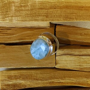 May include: A silver ring featuring a light blue oval-shaped gemstone. The ring is displayed on a wooden surface.