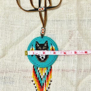 Op de afbeelding: Een turquoise kralenketting met een wolfontwerp en franjes. De ketting heeft een diameter van 7,6 cm en een bruin leren koord.