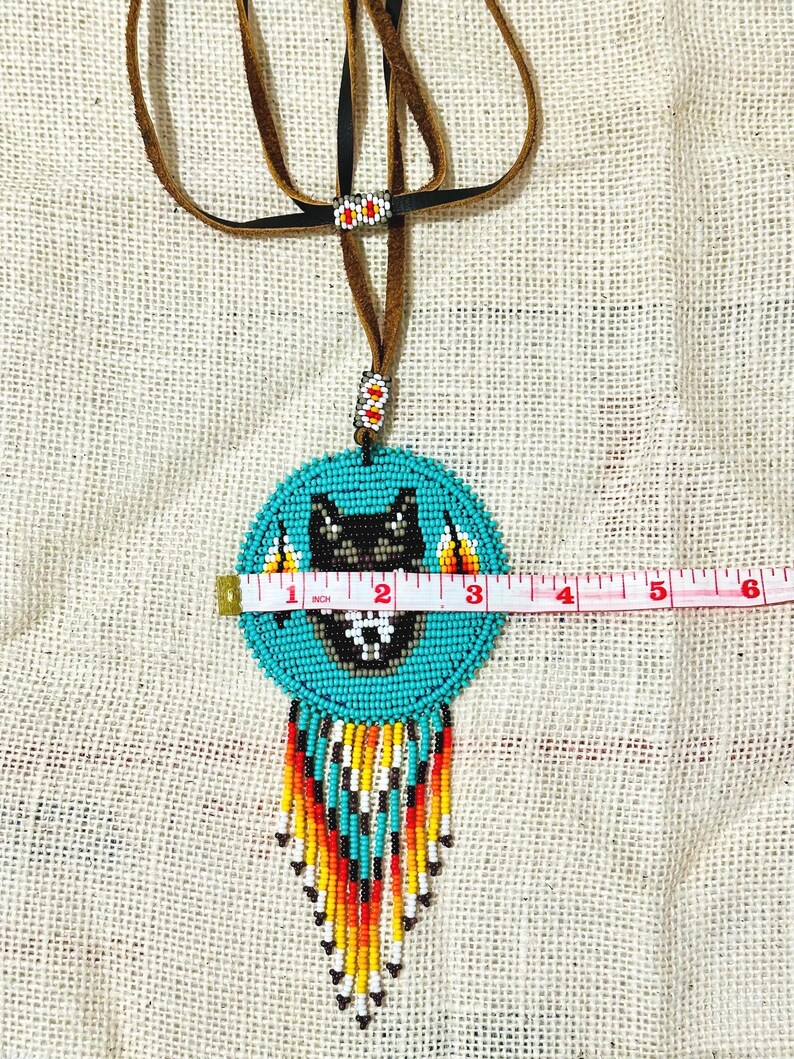 Op de afbeelding: Een turquoise kralenketting met een wolfontwerp en franjes. De ketting heeft een diameter van 7,6 cm en een bruin leren koord.
