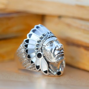 Puede incluir: Un anillo de plata con la cabeza de un jefe nativo americano detallada, que lleva un tocado con plumas negras y blancas y detalles de concha.