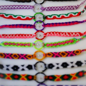 May include: A collection of colorful friendship bracelets with dreamcatcher charms. The bracelets feature various woven patterns in shades of brown, purple, green, red, pink, orange, black, and yellow. Each bracelet has a small, round dreamcatcher charm.