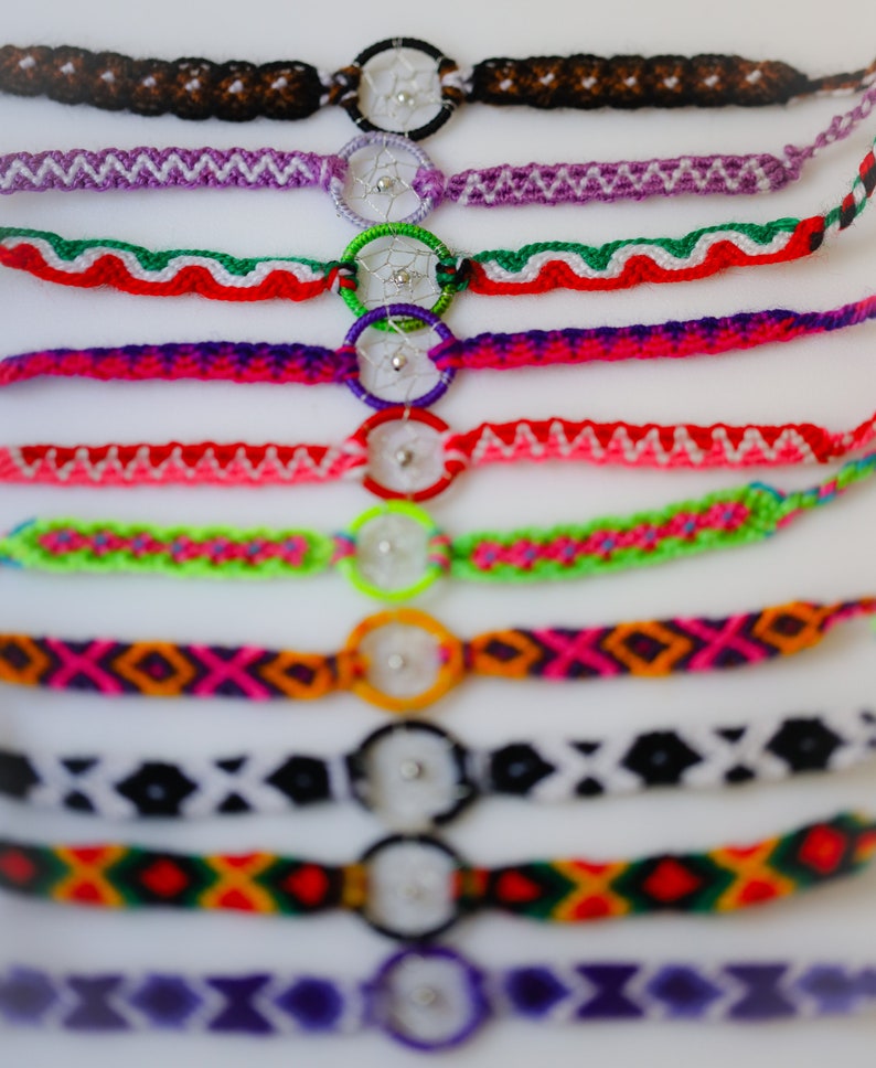 May include: A collection of colorful friendship bracelets with dreamcatcher charms. The bracelets feature various woven patterns in shades of brown, purple, green, red, pink, orange, black, and yellow. Each bracelet has a small, round dreamcatcher charm.