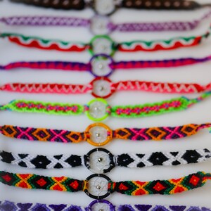 May include: A collection of colorful friendship bracelets with dreamcatcher charms. The bracelets feature various woven patterns in shades of purple, pink, green, orange, black, and white. Each bracelet has a small, circular dreamcatcher charm in the center.