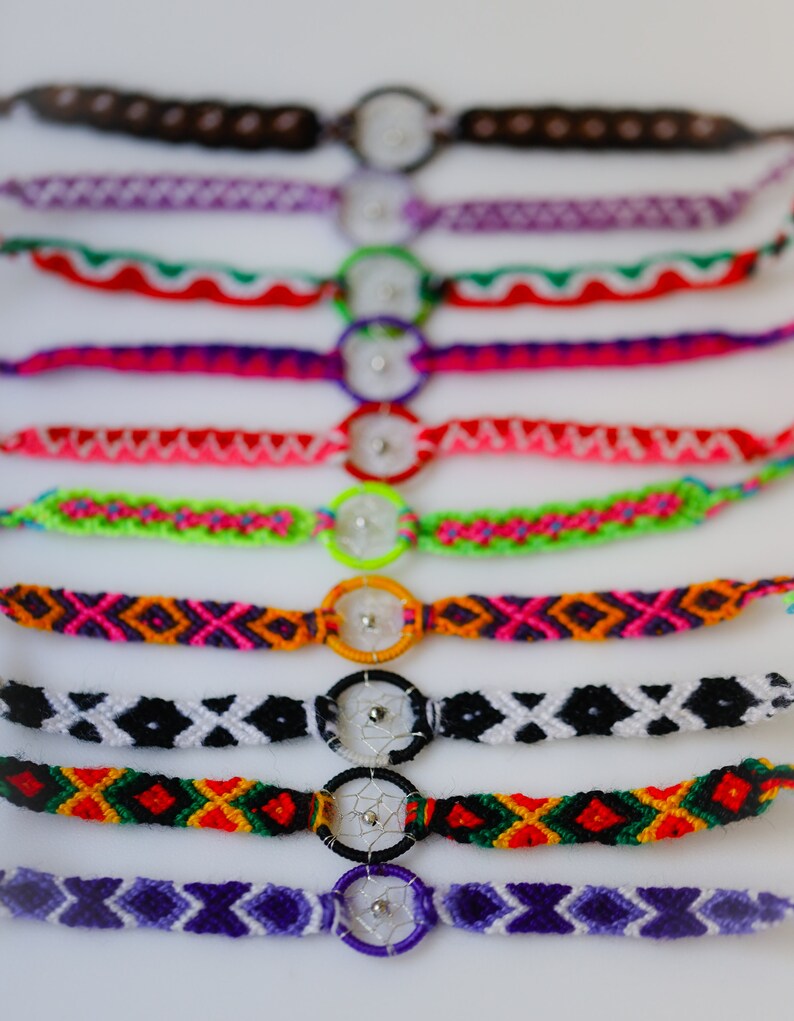 May include: A collection of colorful friendship bracelets with dreamcatcher charms. The bracelets feature various woven patterns in shades of purple, pink, green, orange, black, and white. Each bracelet has a small, circular dreamcatcher charm in the center.