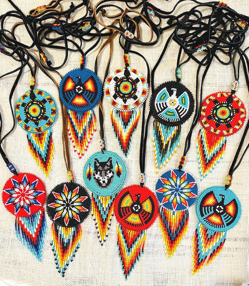 Op de afbeelding: Een verzameling van 12 kralen Native American dreamcatchers met kleurrijke geometrische patronen en franjes. De dreamcatchers zijn geregen aan zwarte leren koorden en hebben verschillende ontwerpen, waaronder een wolf, een vogel en een ster.