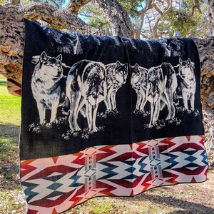 May include: A black and white woven blanket with a pattern of wolves and a geometric design. The blanket is draped over a tree branch.