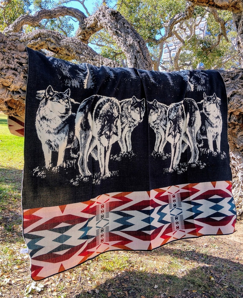May include: A black and white woven blanket with a pattern of wolves and a geometric design. The blanket is draped over a tree branch.