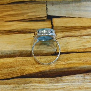 May include: A silver ring featuring a large, oval-shaped, light blue gemstone. The ring is displayed on a wooden surface.