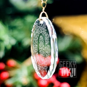 May include: A clear glass ornament with a detailed snowflake design hanging from a gold chain. The ornament is hanging in front of a blurred background of green foliage and red berries.