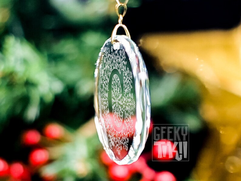 May include: A clear glass ornament with a detailed snowflake design hanging from a gold chain. The ornament is hanging in front of a blurred background of green foliage and red berries.
