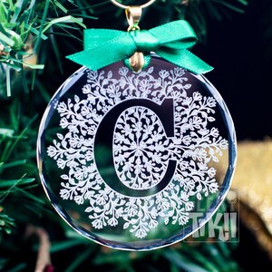 May include: A clear glass Christmas ornament with a white snowflake design and the letter "C" in the center. The ornament is hanging from a green ribbon.