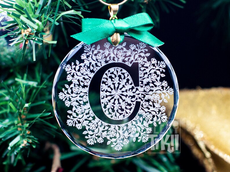 May include: A clear glass Christmas ornament with a white snowflake design and the letter "C" in the center. The ornament is hanging from a green ribbon.