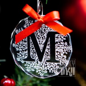 May include: A clear glass Christmas ornament with a snowflake design and the letter "M" in black. The ornament is decorated with a red bow.