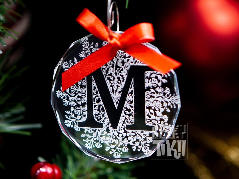 May include: A clear glass Christmas ornament with a snowflake design and the letter "M" in black. The ornament is decorated with a red bow.