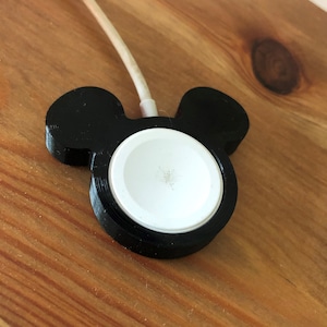 Può includere: Supporto di ricarica nero per Apple Watch a forma di testa di Topolino. La base di ricarica è bianca e il supporto ha un bordo nero. Un cavo di ricarica bianco è collegato al supporto.