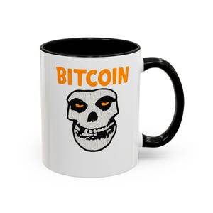 Puede incluir: Taza de cerámica blanca con interior y asa negros. La taza presenta la palabra "BITCOIN" en naranja sobre un gráfico de calavera con ojos naranjas. Una taza de café única para los entusiastas de las criptomonedas.