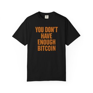 May include: Black t-shirt featuring the orange text "YOU DON'T HAVE ENOUGH BITCOIN". The Bitcoin symbol is included in the text. The shirt has a crew neck and short sleeves.