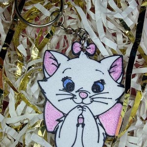 May include: A white cat-shaped keychain with pink accents, blue eyes, and a pink bow. The keychain is attached to a silver metal ring and chain. The cat is smiling with its paws together. The background is gold and white shredded paper.