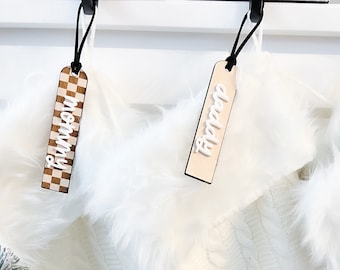 May include: Three white knitted stockings with faux fur trim and personalized wooden tags. The tags read "Mommy", "Fipppop", and "Mattie".