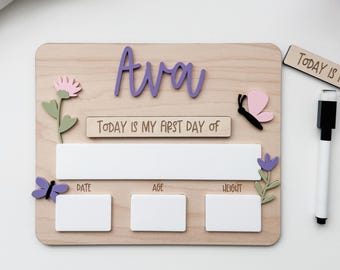 Op de afbeelding: Houten mijlpaalbord met de naam "Ava" in paars schrift. Inclusief ruimte voor datum, leeftijd en gewicht, met decoratieve vlinders en bloemen. Een apart houten bordje leest "Vandaag is mijn laatste dag van". Een zwart-witte marker is inbegrepen.