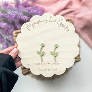 Op de afbeelding: Een houten plaquette met de tekst "Mommy's Love Garden" en de namen "Matteo" en "Avery" met een hartje ertussen. De tekst "loving you since 2019" is ook inbegrepen. Twee groene, bloemvormige houten ornamenten zijn aan de plaquette bevestigd.