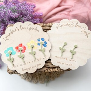 Op de afbeelding: Twee houten borden met de tekst "Grandma's love Garden" en "Mommy's love Garden". Elk bord heeft handafdrukbloemen met namen en data eronder.