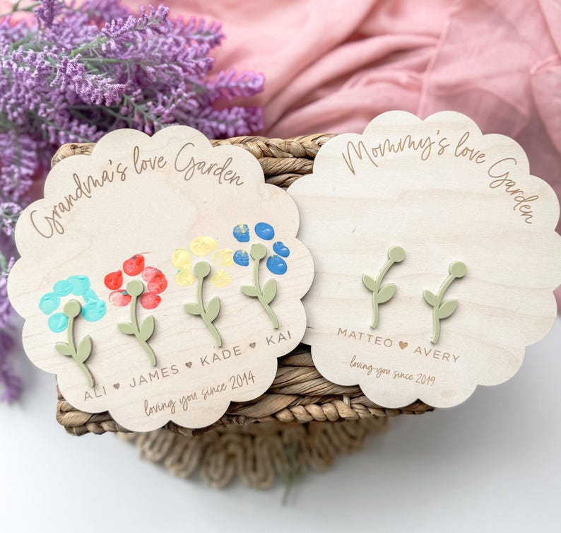 Op de afbeelding: Twee houten borden met de tekst "Grandma's love Garden" en "Mommy's love Garden". Elk bord heeft handafdrukbloemen met namen en data eronder.