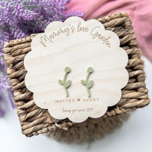Op de afbeelding: Een houten bord met de tekst "Mommy's Love Garden" en de namen "Matteo" en "Avery" met een hartje ertussen. De tekst "loving you since 2019" staat onder de namen. Twee groene bloemoorbellen worden op het bord getoond.