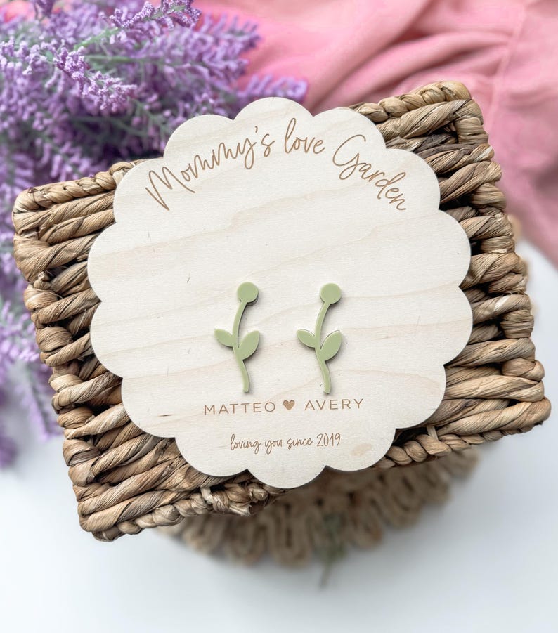Op de afbeelding: Een houten bord met de tekst "Mommy's Love Garden" en de namen "Matteo" en "Avery" met een hartje ertussen. De tekst "loving you since 2019" staat onder de namen. Twee groene bloemoorbellen worden op het bord getoond.