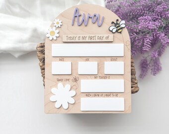 Op de afbeelding: Een houten bord voor de eerste schooldag met de naam "Ava" in paars. Het bord bevat vakken voor datum, leeftijd en gewicht, samen met "Dingen die ik leuk vind" en "Mijn leraar is". Decoratieve elementen zijn een bij en bloemen.