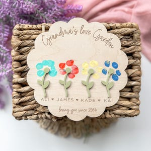 Op de afbeelding: Een houten bord met de tekst "Grandma's Love Garden" en vier handafdrukbloemen in verschillende kleuren. De bloemen zijn gelabeld met de namen Ali, James, Kade en Kai. Het bord heeft ook de tekst "loving you since 2014".