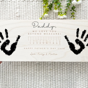 May include: A light-colored wooden plaque with two black handprints and the text "Daddy, WE LOVE YOU BEYOND MEASURE!" A ruler graphic is present, with the text "HAPPY FATHER'S DAY 2024" and the names "Love- Avery & Matteo".