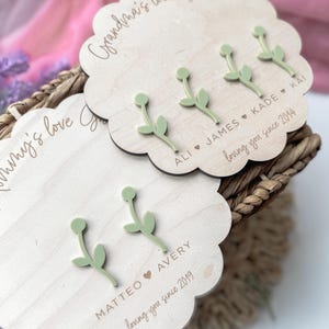 Op de afbeelding: Drie houten plaquettes met de tekst "Grandma's love Garden", "ALI JAMES KADE KAI loving you since 2014" en "MATTEO AVERY loving you since 2019". Elke plaquette heeft een groene houten bloem.