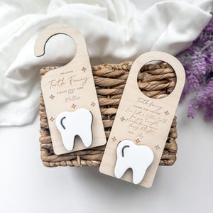 Peut inclure: Deux panneaux de porte en bois sur le thème de la fée des dents. Chacun a une découpe de dent blanche et le texte "Bienvenue Fée des dents" et "Fée des dents Merci de votre visite". Les cintres sont marron clair.