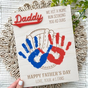May include: Wooden sign with the words "Daddy" in red, "We hit a home run scoring you as ours" and "Happy Father's Day Love, Your #1 Fans". The sign features a baseball glove with red and blue handprints.