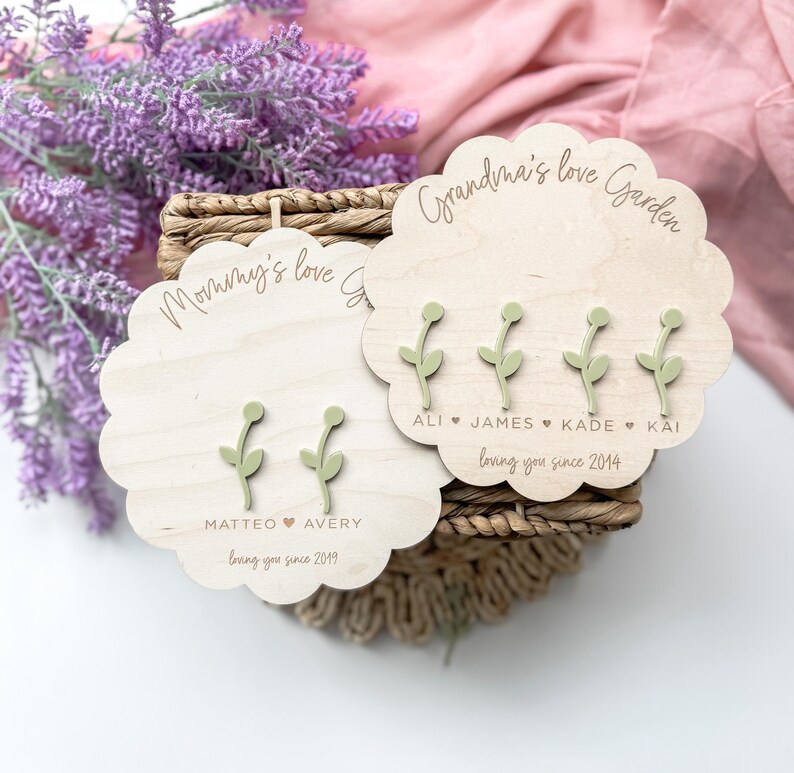 Op de afbeelding: Twee houten plaquettes met de woorden "Mommy's Love Garden" en "Grandma's Love Garden" erop gegraveerd. Elke plaquette heeft een ander aantal groene bloemuitsparingen met namen en data eronder.