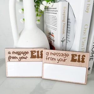 Pode incluir: Dois sinais de madeira com fundo branco e o texto "a message from your Elf" em letras marrons. Os sinais são perfeitos para deixar mensagens dos duendes do Papai Noel.