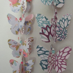 May include: A string of paper butterflies with floral patterns in shades of pink, blue, and green. The butterflies are strung together with silver thread and small beads.