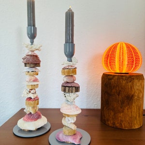 May include: Two gray candle holders with gray candles, each stacked with a variety of seashells and driftwood. The candle holders are on a wooden table next to a wooden base with a round orange lampshade.