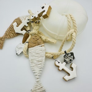 Könnte beinhalten: Dekorative Artikel zum Thema Nautik, darunter eine weiße Rettungsboje, Seil und seilumwickelte Fisch- und Meerjungfrauenformen. Holzanker mit weißem Emaille sind am Seil befestigt. Die Artikel sind in Weiß-, Beige- und Brauntönen gehalten.