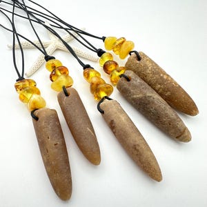 Op de afbeelding: Vier kettingen met langwerpige, taps toelopende bruine stenen aan zwarte koorden. Elke hanger is versierd met drie doorschijnende, goudgele barnstenen kralen boven de steen. De stenen hebben een gladde, verweerde textuur.