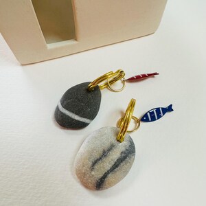 Può includere: Orecchini con pietre ovali e cerchi dorati. Uno ha una pietra grigio scuro con una striscia bianca, l'altro una pietra grigio chiaro con strisce grigio scuro. Ognuno ha un piccolo ciondolo a forma di pesce, uno rosso e uno blu.