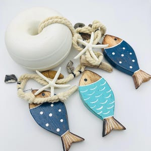 Könnte beinhalten: Dekoration im maritimen Stil. Weiße Boje, Seil, Seesterne und drei Holzfische. Die Fische sind blau und türkis bemalt mit weißen Punkt- und Schuppendetails sowie Flossen aus Naturholz. Küsten-Dekoration.