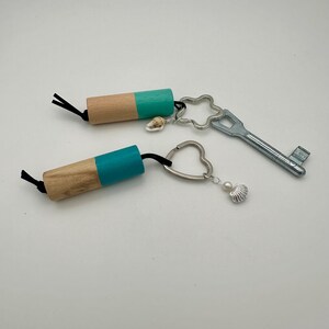 Può includere: Due portachiavi con cilindri di legno dipinti di verde acqua, attaccati a cordoncini neri. Uno ha un ciondolo a forma di chiave d'argento, l'altro un ciondolo a forma di cuore. Entrambi hanno accenti di conchiglie e perle. È presente anche una chiave d'argento.