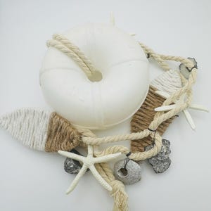 Könnte beinhalten: Dekorative nautische Komposition mit einem weißen Rettungsring, Seil, Seesternen und kleinen Steinen. Das Seil ist um den Rettungsring und andere Elemente gewickelt. Die Farbpalette umfasst Weiß, Beige und Braun.