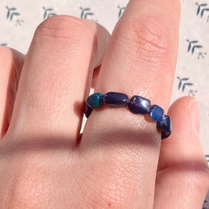 Könnte beinhalten: Ein Ring aus dunkelblauen und grünen Steinen. Der Ring befindet sich an einem Finger.