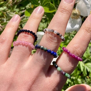 May include: Six beaded rings made of different gemstones. The rings are stacked on a hand, with the black ring on the index finger, the pink ring on the middle finger, the blue ring on the ring finger, the red ring on the pinky finger, and the green ring on the thumb. The rings are all made of small, round beads.