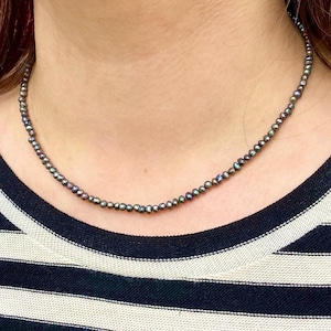 Può includere: Una collana semplice realizzata con piccole perle nere iridescenti. La collana è indossata da una persona con i capelli scuri e una camicia a righe.